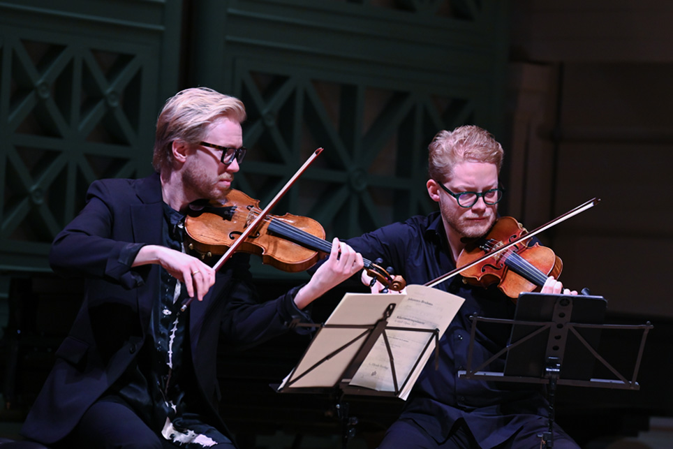 Two male students, wearing glasses, playing the violin in a string quartet, on a dark stage.